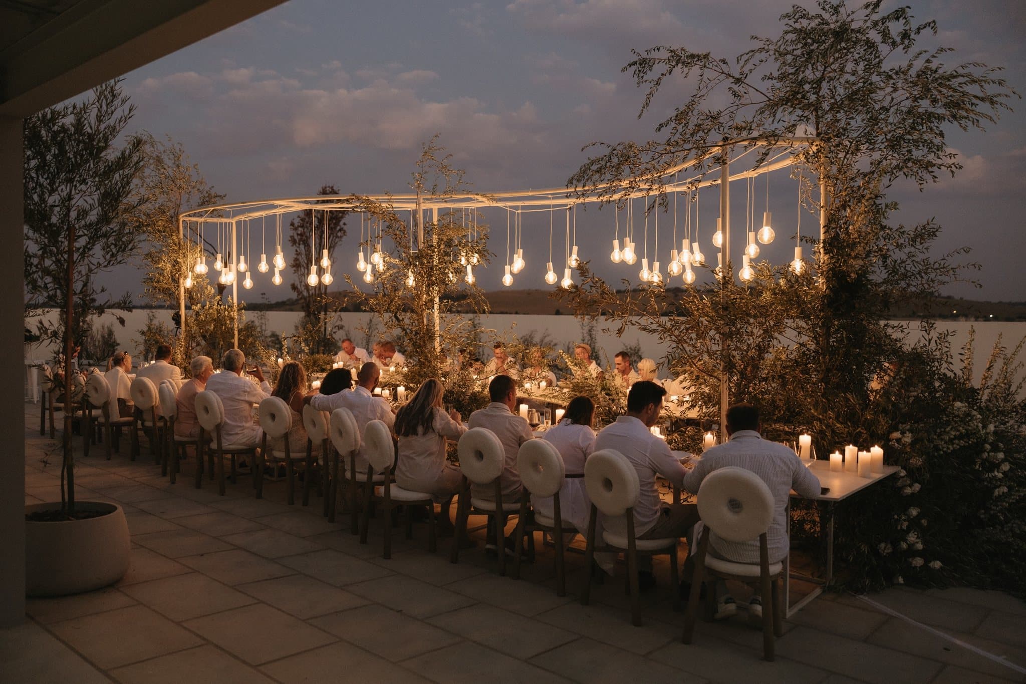 A candlelit dinner on the terrace at dusk, pendant lights strung over the long table above the water